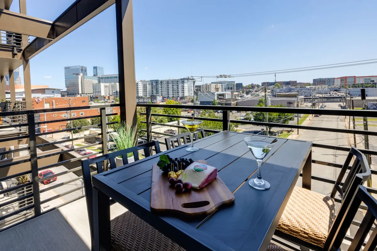 An outdoor dining table set for a social gathering, complete with a glass of martini, a cheese platter, and lush green plants, all set against a backdrop of a glowing city skyline at dusk.