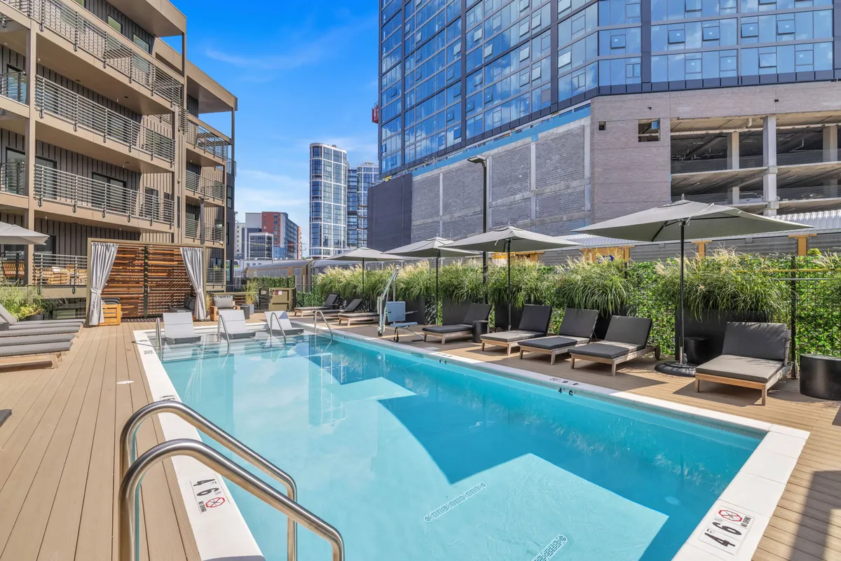 A sparkling outdoor swimming pool is surrounded by comfortable lounge chairs, lush greenery, and the facades of modern buildings. The pool area is sunlit, with evening ambiance hinting at sunset views, and there’s space for socializing, relaxing, and sunbathing.