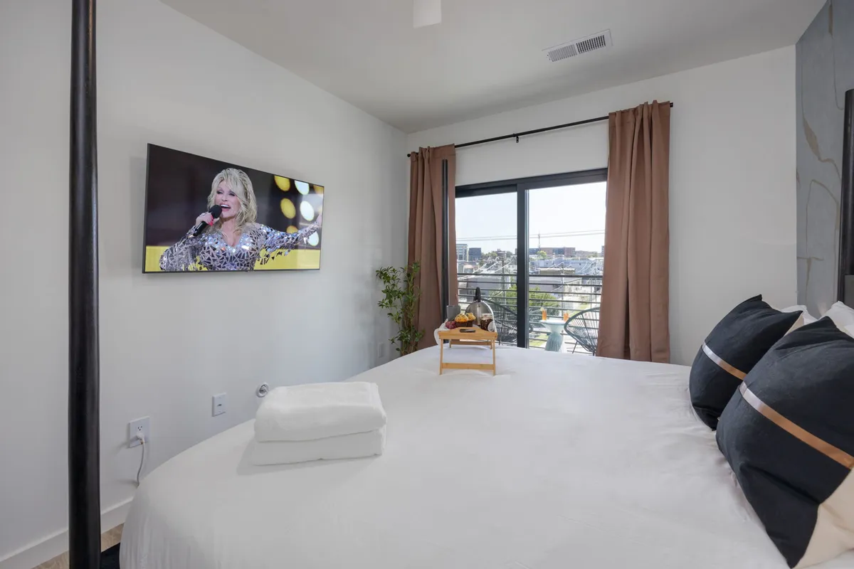 A spacious bedroom with a large bed adorned with decorative pillows, a television screen mounted on the wall, a window letting in natural light, and a green plant adding a touch of freshness. The ambiance is cozy and inviting, perfect for unwinding.