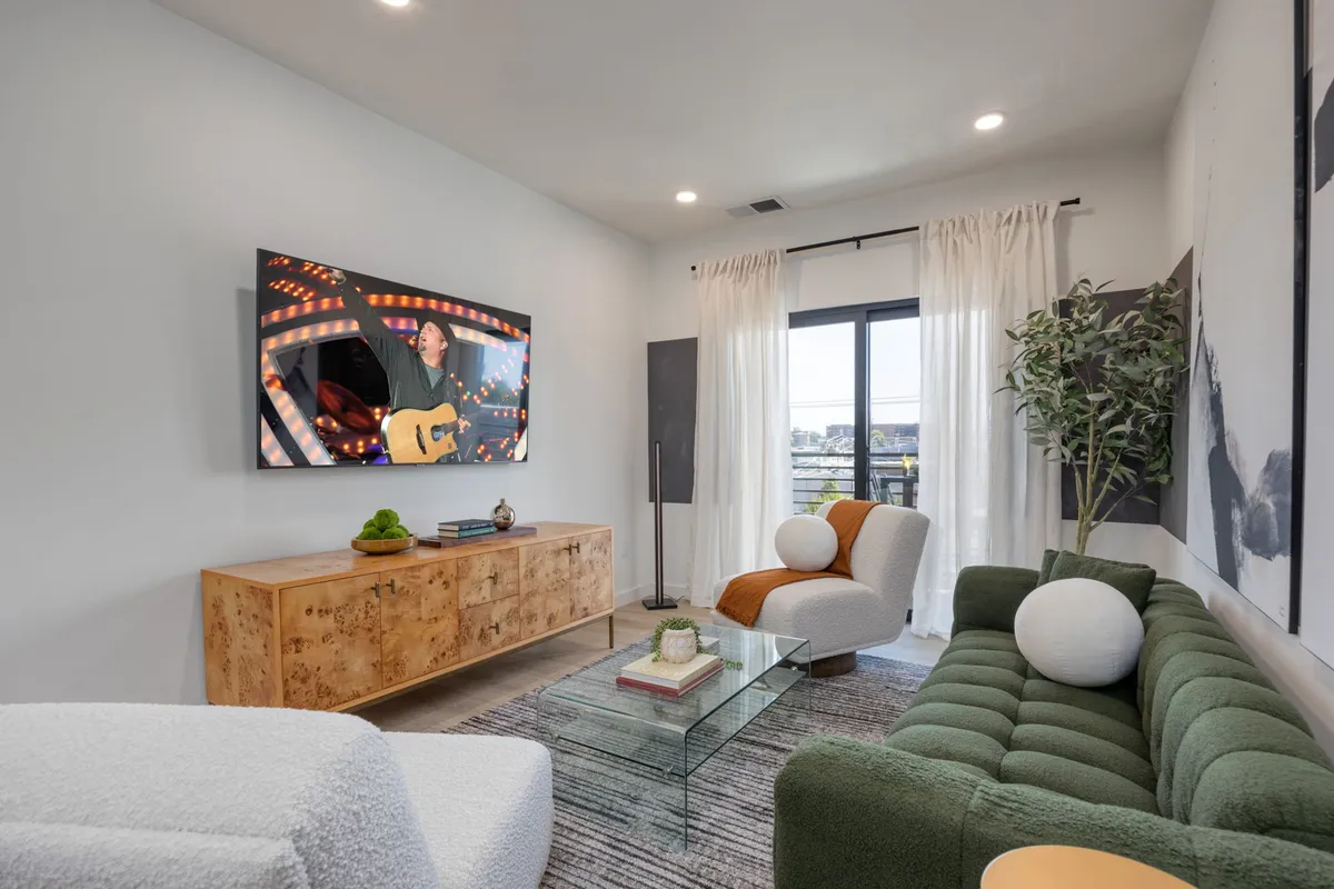 The image showcases a chic, modern living room with a plush green couch, a stylish wooden sideboard, a sleek glass coffee table, and a large television displaying a live concert performance. The space is filled with natural light and decorated with contemporary art, creating a vibrant yet comfortable atmosphere perfect for relaxing or entertaining.