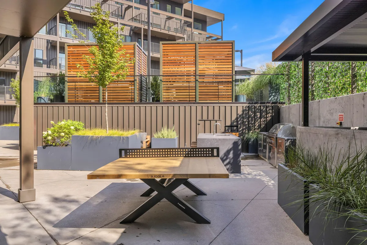 An inviting outdoor dining and grilling area with a wooden table, lush green plants, a modern grilling station, and wooden privacy screens. The space feels casual and welcoming, designed for social gatherings, outdoor meals, or impromptu celebrations with neighbors.