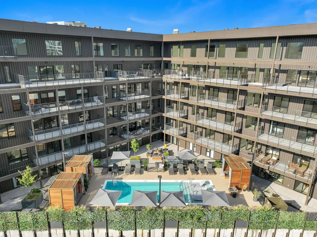 A lively poolside scene featuring a sparkling swimming pool surrounded by lounge chairs, stylish outdoor lounge seating, lush greenery, and glimpses of apartment balconies—inviting for gatherings both large and small.