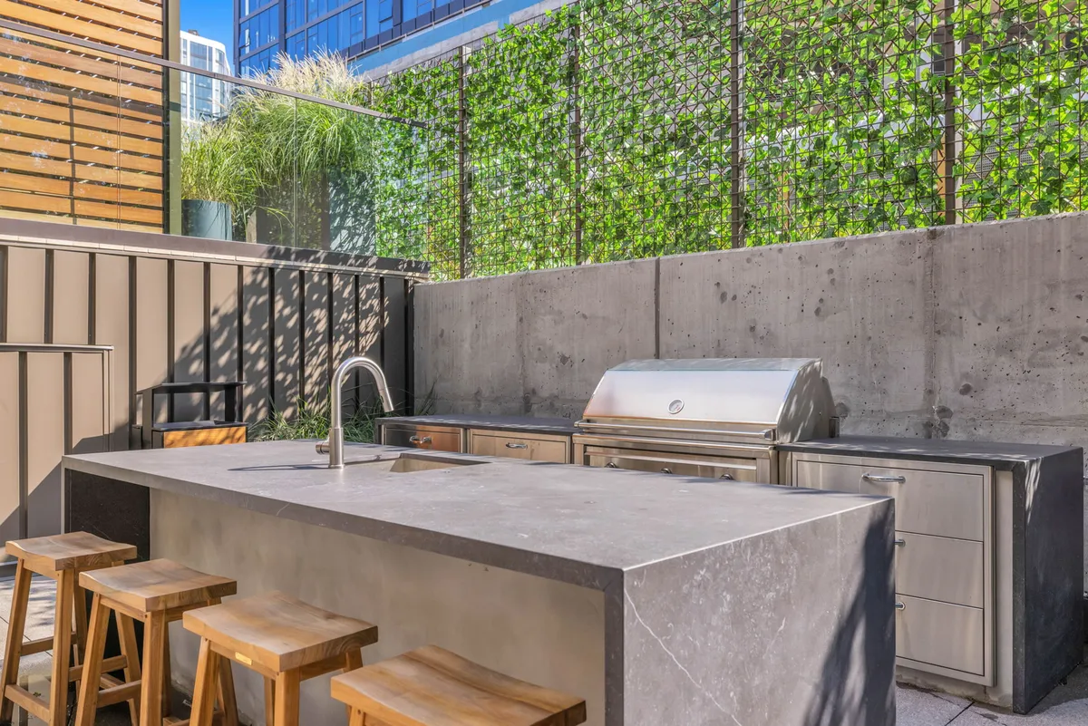 An inviting outdoor kitchen island features a built-in grill, bar stools for seating, and a lush green wall backdrop. The space is designed for al fresco cooking, dining, and gathering with friends and family during sunset or evening events.