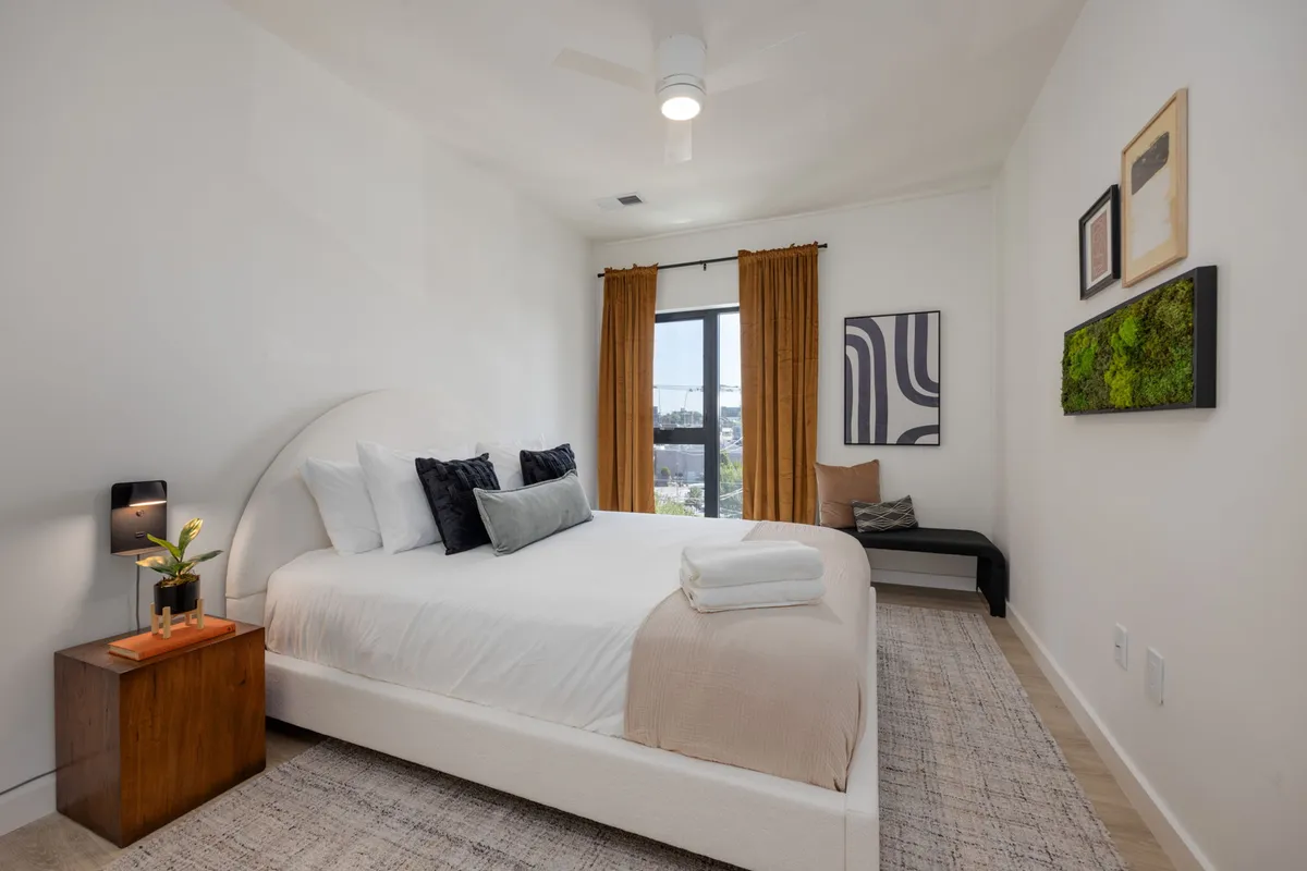 A serene bedroom with a comfortable bed, curtains framing a window, art pieces above the headboard, and a side table with a plant. Soft morning light creates a peaceful vibe ideal for rest and relaxation.